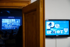 Wystawa: Powroty - Wejście do Galerii TCK, na pierwszym planie telewizor z grafiką wystawy, na drugim planie projekcja filmu.