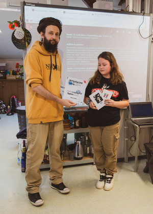 Artystyczne Ferie - tydzień II - Na fotografii uwieczniono grupę uczestników – dzieci, młodzież oraz dorosłych – zaangażowanych w warsztaty tworzenia Artbooków. Widoczne są różnorodne narzędzia, w tym prasa drukarska, z których korzystają pod czujnym okiem prowadzących.