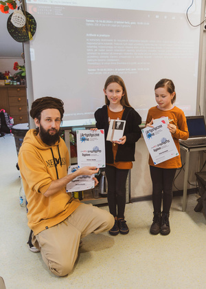 Artystyczne Ferie - tydzień II - Na fotografii uwieczniono grupę uczestników – dzieci, młodzież oraz dorosłych – zaangażowanych w warsztaty tworzenia Artbooków. Widoczne są różnorodne narzędzia, w tym prasa drukarska, z których korzystają pod czujnym okiem prowadzących.
