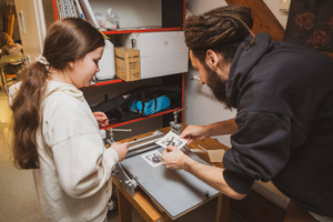 Artystyczne Ferie - tydzień II - Na fotografii uwieczniono grupę uczestników – dzieci, młodzież oraz dorosłych – zaangażowanych w warsztaty tworzenia Artbooków. Widoczne są różnorodne narzędzia, w tym prasa drukarska, z których korzystają pod czujnym okiem prowadzących.