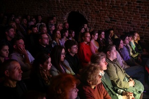 Koncert: Pracownia piosenki prezentuje - Niepozorny zaścianek - Zdjęcie pokazuje widownię w Piwnicach TCK podczas koncertu. Widzowie, siedzący na krzesłach obserwują występ. Na zdjęciu widać różnorodność publiczności, od młodych po starszych słuchaczy. Sala jest delikatnie oświetlona.