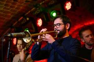 Koncert jazzowy: Tomasz Chyła Quintet - Zdjęcie sceny z koncertu Tomasz Chyła Quintet, pokazujące zaangażowanych muzyków. Tomasz Chyła gra na skrzypcach, w tle pozostali muzycy na różnych instrumentach. Scena jest dynamicznie oświetlona, co dodaje energii występowi.