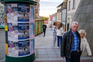 Wystawa: Zrozumie życie - Słup ogłoszeniowy z plakatami zapowiadającymi wystawę i mijający go pierwsi goście wernisażu wystawy.
